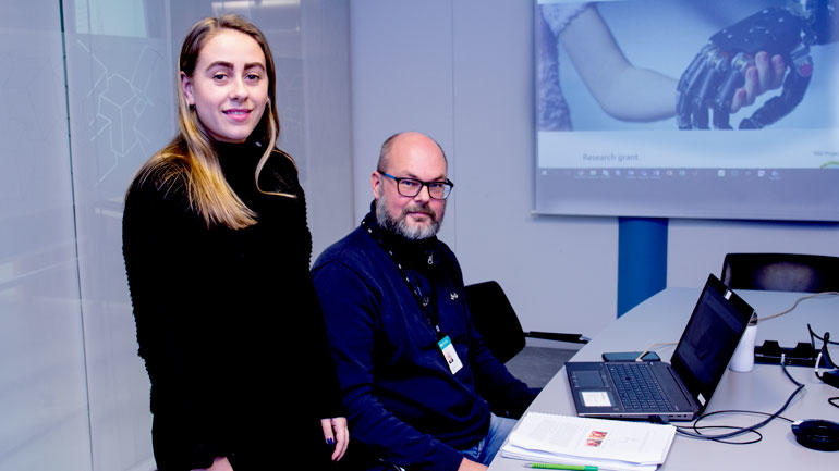 Student Hanna Borch og prosjektleder Lars-Cyril Blystad foran bilde av armprotesen og ser rett i kamera 