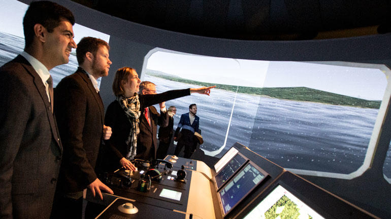 Statsråd Iselin Nybø tar styringen på det maritime simulatorlaboratorium på USN. Til venstre professor Salman Nazir og bachelorstudent i nautiske operasjoner, Adrian Andersen.