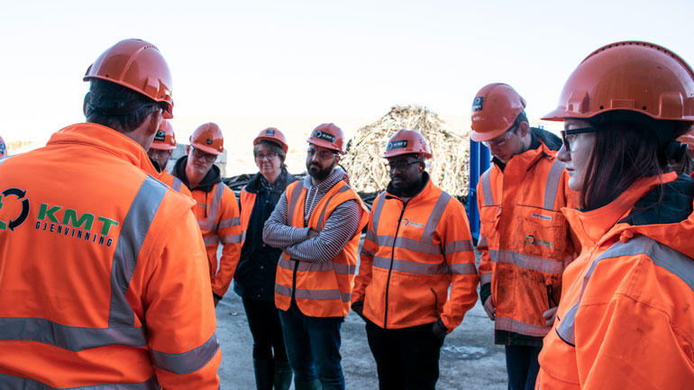 Ingeniører i oransje arbeidstøy og med oransje hjelmer på hodet står på en rekke på en industriplass og hører på en mannlig ingeniør som snakker. Illustrasjonsbilde