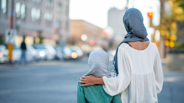 Mor og datter iført hijab poserer med ryggen mot kamera foran et urbant miljø. Foto