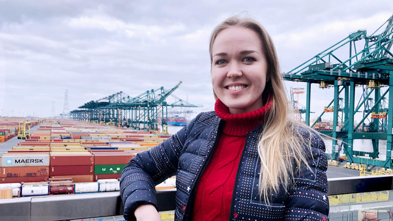 Mariia i rød høyhalsa genser og dunjakke, langt, lyst hår smiler til kamera foran et havnemiljø. 