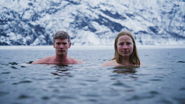 ein mann og ei kvinne badar vinterstid med høge fjell i bakgrunnen. Foto