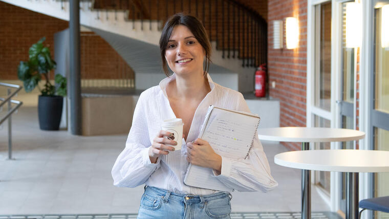 IT-student Maria Elena Cata smiler og holder en kaffekopp og en notatblokk. Foto.